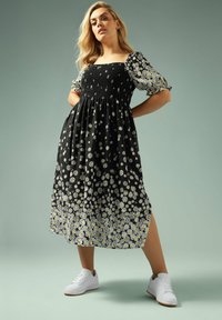 Svart blommig klänning med smockad bodice, puffärmar och ankellängd. Matchad med vita sneakers och en enfärgad bakgrund.