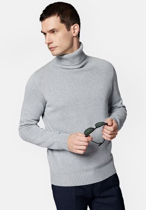 Szary sweter z golfem, wykonany z miękkiej przędzy dzianinowej, z prążkowanymi mankietami i dołem. Model trzyma czarne okulary przeciwsłoneczne, na jednolitym tle.