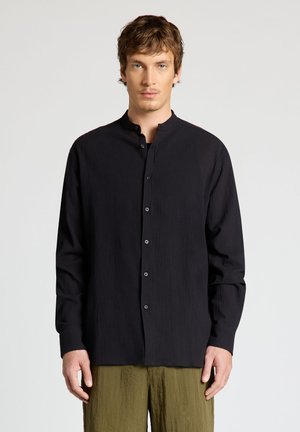 Jeune homme aux cheveux châtain clair porte une chemise noire à col mandarin boutonnée et un pantalon vert olive, debout devant un fond clair uni.