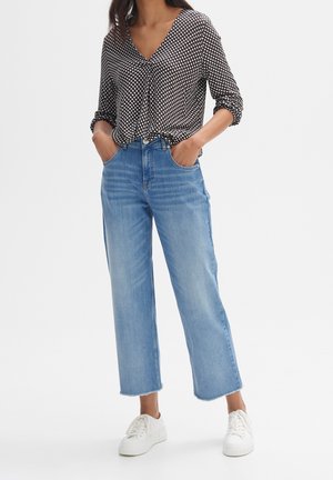 Mujer con blusa de estampado blanco y negro, vaqueros cortos azul claro con dobladillos sin rematar y deportivas blancas con cordones, manos en los bolsillos.