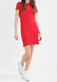 Vestido polo rojo con mangas cortas, una tapeta de tres botones y aberturas laterales. Combinado con zapatillas altas blancas. Tejido suave y ajustado.