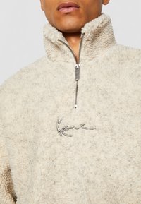 Beiger Fleece-Pullover mit hohem Kragen, halbem Reißverschluss und strukturiertem Obermaterial. Verfügt über ein kontrastierendes silbernes Logo auf der Brust.