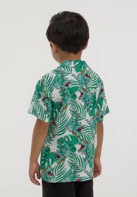 Camicetta a maniche corte con stampa di un dinosauro verde su sfondo bianco con motivi di foglie tropicali. Design leggero e informale.
