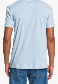 Persona vestida con una camiseta de manga corta azul claro y pantalones vaqueros azules, mostrada de espaldas sobre un fondo blanco.