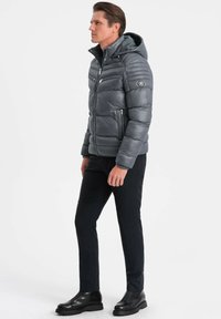 Giubbotto in piuma grigio con cappuccio, dotato di tasche con zip e cuciture orizzontali. Indossato con pantaloni scuri e stivaletti neri.