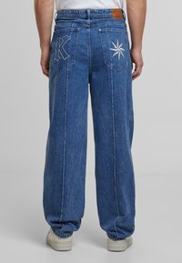 Siniset denim leveälahkeiset farkut, joissa on korkea vyötärö. Takataskuissa on kirjailtu "K"- ja tähti-muotoilu sekä hienovarainen pinta.
