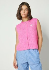 Ilma varrukatega roosa kootud vest pehme tekstuuriga, mille esiosal on nupud ja väike logo silt. Sobib triibuliste siniste pükstega.