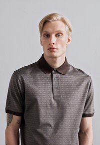 Brūna polo krekla ar atkārtotu "Emporio Armani" logo rakstu, trīs pogu aizdari un tumši brūnu apkakli ar īsām piedurknēm.