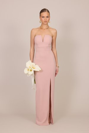 Femme en robe rose poudré sans bretelles avec une fente devant, tenant des callas blancs, portant des boucles d'oreilles et un collier en or, debout devant un fond uni.