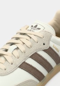 Primer plano de una zapatilla Adidas Samba con cordones beige, franjas marrones y texto "SAMBA" en la parte superior de cuero blanco.