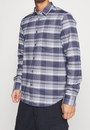 Homme portant une chemise à carreaux bleue et blanche à manches longues boutonnée et un pantalon foncé, debout devant un fond uni.