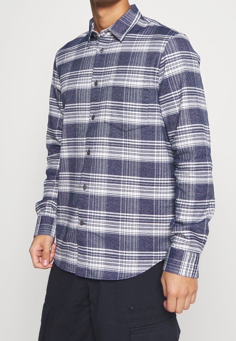 Homme portant une chemise à carreaux bleue et blanche à manches longues boutonnée et un pantalon foncé, debout devant un fond uni.