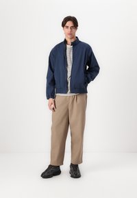 Ναυτικό μπλε ελαφρύ μπουφάν με ελαστικούς καφέδες, συνδυασμένο με ριγέ πουκάμισο και μπεζ παντελόνι. Το μοντέλο φορά μαύρα αθλητικά παπούτσια. Απλό, casual ντύσιμο.