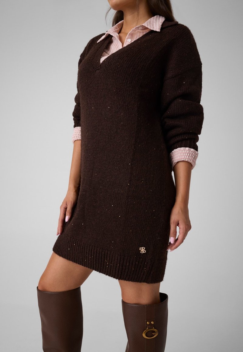 Femme portant une robe pull en maille marron foncé par-dessus une chemise à col rose clair et des bottes en cuir marron jusqu’aux genoux.