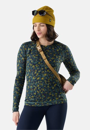 Mujer con un gorro de punto amarillo con gafas de sol, una camisa verde floral de manga larga, pantalones azul marino y una correa de bolso cruzado marrón claro.