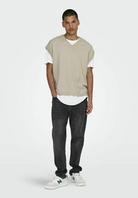 Beige übergroßer Strickwesten mit V-Ausschnitt über einem weißen T-Shirt, kombiniert mit dunklen Jeans und weißen Sneakers mit grünen Akzenten.