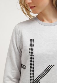 Sudadera gris con un patrón de puntos negros texturizados en la parte delantera. Cuello redondo y mangas largas, hecha de un tejido suave y elástico.