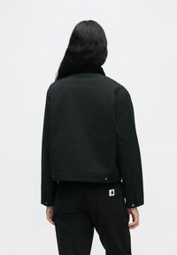 Άτομο με μακριά μαύρα μαλλιά, φορώντας μαύρο jacket και μαύρο παντελόνι, στέκεται γυρισμένο από την κάμερα με φόντο λευκό.