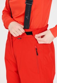 Rode broek met verstelbare zwarte bretels en een ritszak aan de zijkant. Het materiaal lijkt glad met een lichte glans.