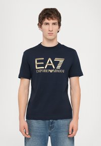 Tamsiai mėlyna medvilnės marškin�ėliai su apvaliu iškirpimu ir trumpomis rankovėmis. Priekyje pateikiama auksinė grafika su logotipu "EA7 Emporio Armani".