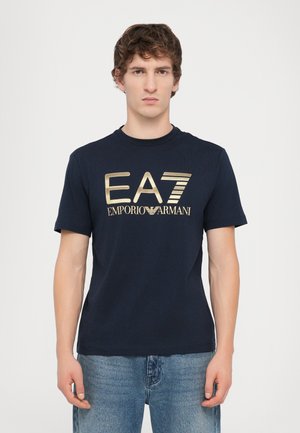 T-shirt en coton bleu marine avec un col rond et des manches courtes. Présente un logo graphique doré "EA7 Emporio Armani" sur le devant.