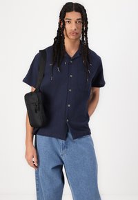 Navy short-sleeve button-up shirt with a textured fabric, left chest pocket, paired with light blue loose jeans and a small black crossbody bag.
