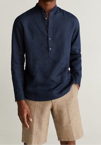 Camicia di lino blu navy con colletto alla coreana, maniche lunghe e bottoni. Indossata con pantaloni corti di lino beige chiaro.