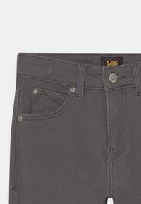 Vaqueros de mezclilla gris con un diseño clásico de cinco bolsillos, herrajes de metal y una etiqueta de marca visible en la cintura. Acabado en tela texturizada.