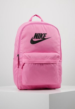 Lyserød Nike rygsæk med forreste lynlåslomme og sort Nike-logo, vist på en hvid piedestal mod en grå baggrund.