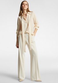 Crèmekleurige blouse met zijden uitstraling en een strik, gecombineerd met hoge, wijde crèmekleurige broeken. Puntige, naakte hakken maken de outfit compleet.