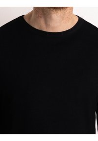 Torso maschile che indossa un maglione nero a collo rotondo, che mostra il collo e una parziale barba su uno sfondo chiaro e uniforme.