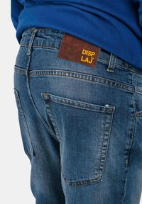 Jeans in denim blu scuro con una finitura texturizzata. Presentano tasche standard e una patch in pelle marrone etichettata "DISP LAJ." Accenti di cucitura minimal.
