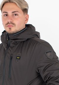 Giacca con cappuccio marrone scuro dal texture liscia e lucida. Presenta una tasca sul petto con zip e un distintivo sulla manica sinistra.