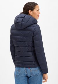 Frau im Seitenprofil, trägt eine marineblaue gesteppte Kapuzenjacke und blaue Jeans vor einem schlichten weißen Hintergrund.