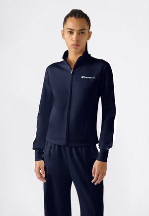 Frau mit geflochtenem Haar, die eine marineblaue Champion-Trainingsjacke mit Reißverschluss und passende Hosen trägt, vor einem schlichten weißen Hintergrund steht.