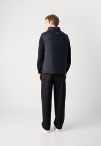 Gilet imbottito blu navy con alto colletto, sezioni divise e toppa con logo sul retro, abbinato a pantaloni neri larghi e scarpe bianche.