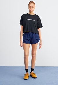 Črna skrajšana majica s belim logotipom, mornarsko modri kratki hlačnici s črtami, črne nogavice in rumeni teniski čevlji. Stoči na modri podlagi ob svetli ozadju.