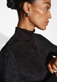 Top nero a collo alto con una finitura testurizzata e luccicante. Il modello indossa un orecchino a forma ovale in argento. Focus sullo scollo e sui dettagli del tessuto.