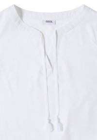 Blouse blanche avec col rond, fente avant et deux liens à pompons suspendus, étiquetée avec la marque "CECIL" à l'intérieur du col.