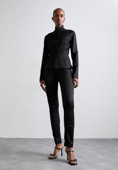 Blazer noir ajusté avec col haut et fermeture à boutons, associé à un pantalon noir sur mesure et des talons noirs à lanières. Texture de tissu lisse.