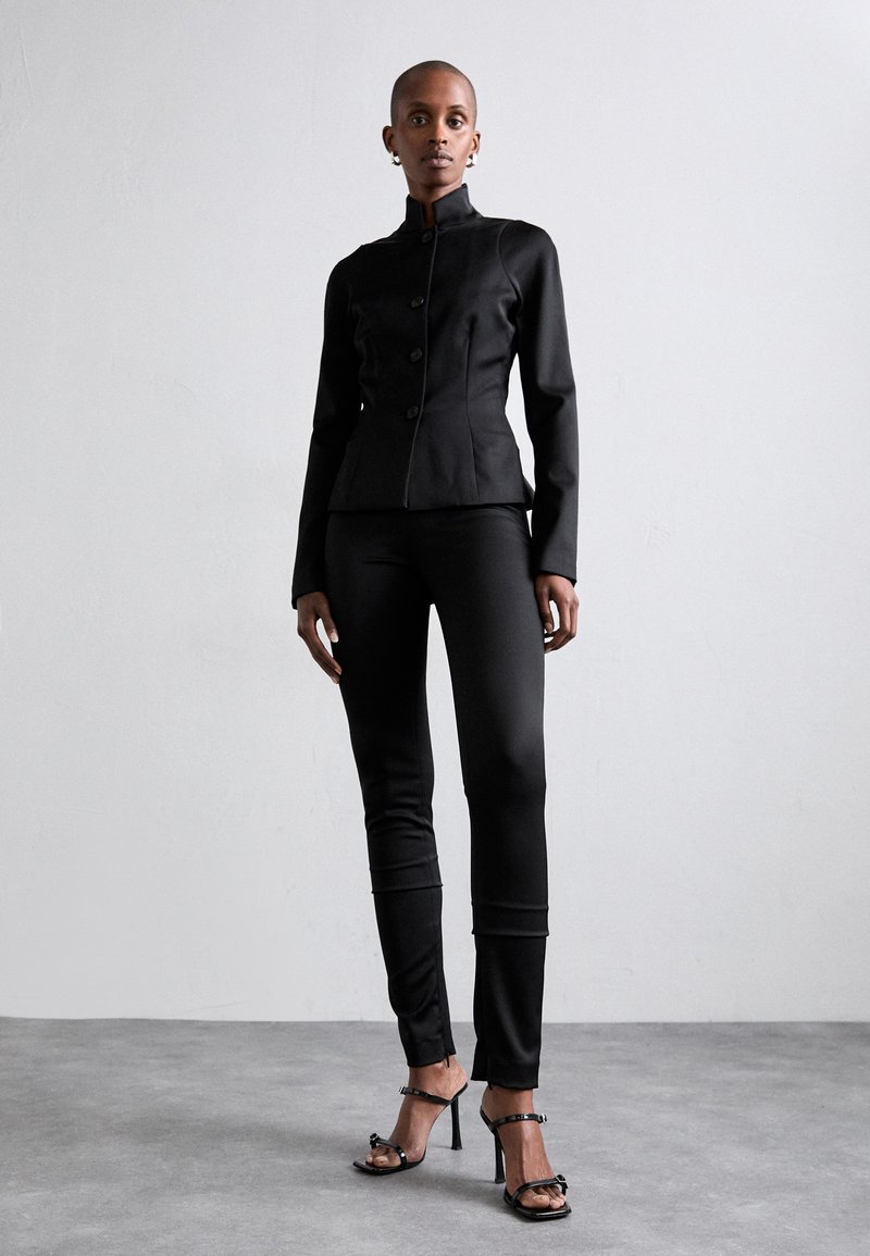 Fitted black blazer with high collar and button closure, paired with black tailored pants and strappy black heels. Smooth fabric texture.