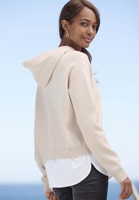 Beige hoodie met geribbelde onderrand, gecombineerd met een wit shirt eronder en donkere jeans. De stof lijkt zacht en glad.