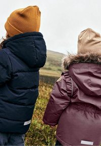 Dvě děti stojící zády k fotoaparátu, oblečené v námořnické prošívané bundě a vínovém kabátu s kožešinovou kapucí.