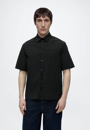 Man die een zwart overhemd met korte mouwen en knopen en donkerblauwe jeans draagt, staande tegen een effen witte achtergrond.