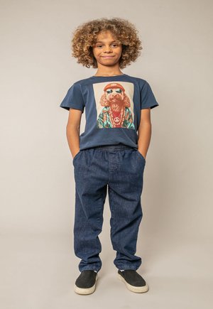 Enfant aux cheveux bouclés, les mains dans les poches, portant un t-shirt bleu marine avec un chien en lunettes de soleil et chapeau, un pantalon en denim ample, et des baskets noires sans lacets.