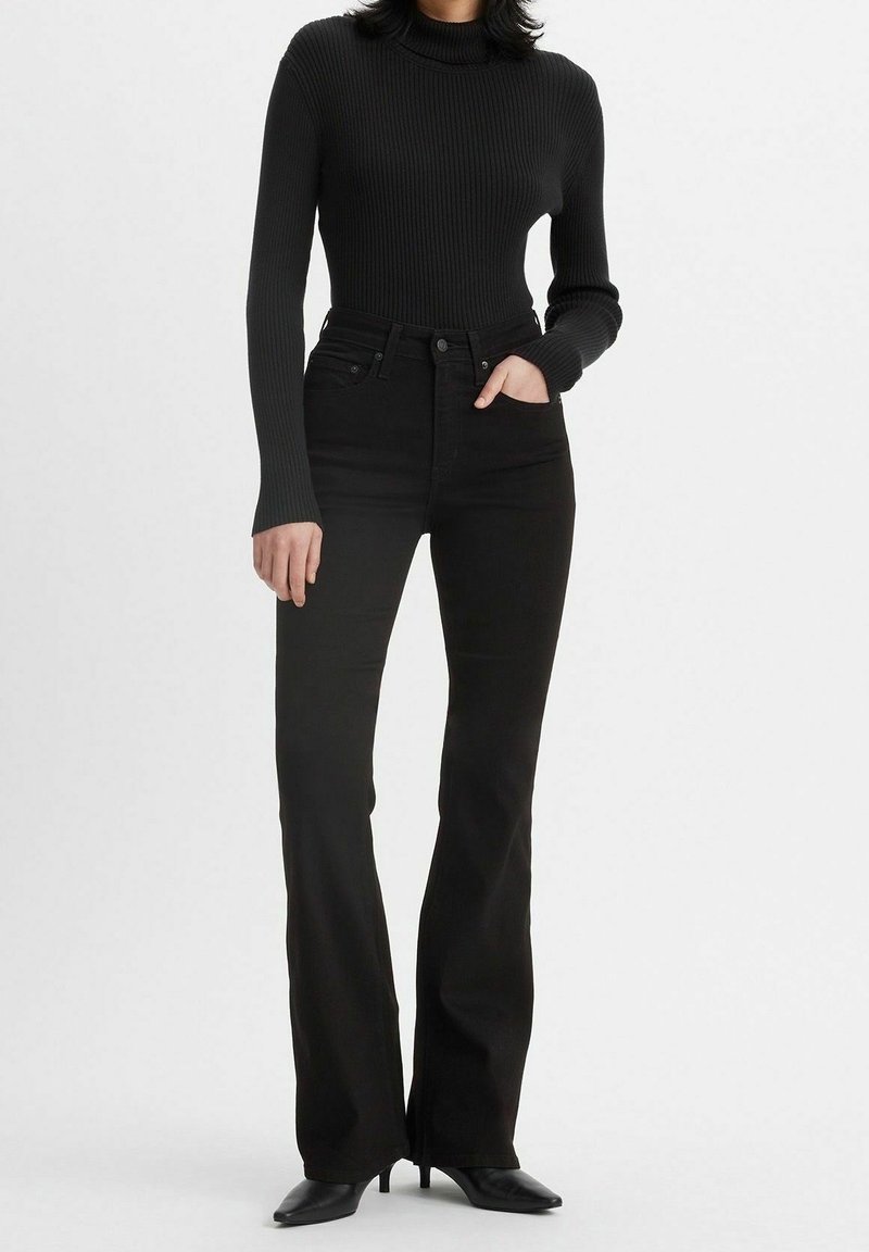 Femme portant un jean évasé noir taille haute, un pull à col roulé en côte noir et des chaussures à talons pointues noires, main dans la poche.