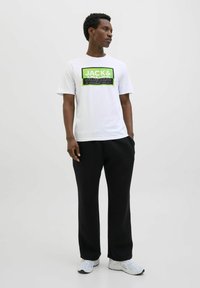 Weißes Baumwoll-T-Shirt mit einem schwarzen und grünen grafischen Logo; kombiniert mit schwarzen Jogginghosen und weißen Sportschuhen. Schlichtes Design.