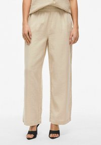 Pantalons larges beiges en tissu texturé, dotés d'une taille élastique et de fentes sur les côtés, associés à des talons ouverts noirs.
