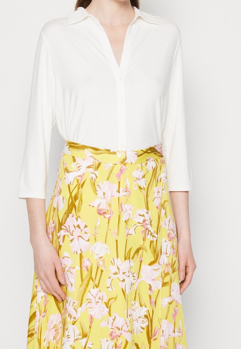 Femme portant une chemise blanche boutonnée avec des manches trois-quarts et une jupe fleurie jaune avec des fleurs roses et marron.