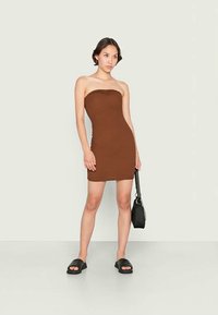 Vestido bodycon castanho com design sem alças, apresentando uma silhueta ajustada. Combinado com sandálias pretas tipo slide e uma pequena mala.